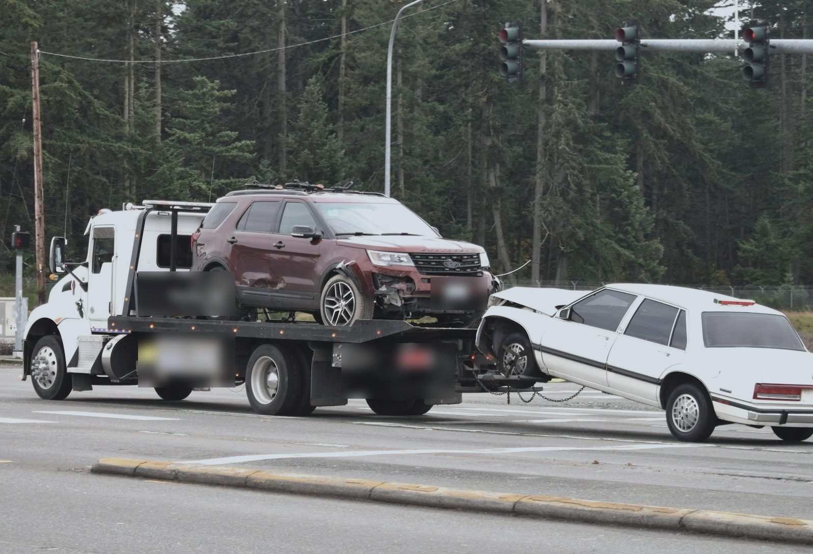 A flatbed tow truck at an intersection carrying an SUV on its deck and pulling a white sedan behind