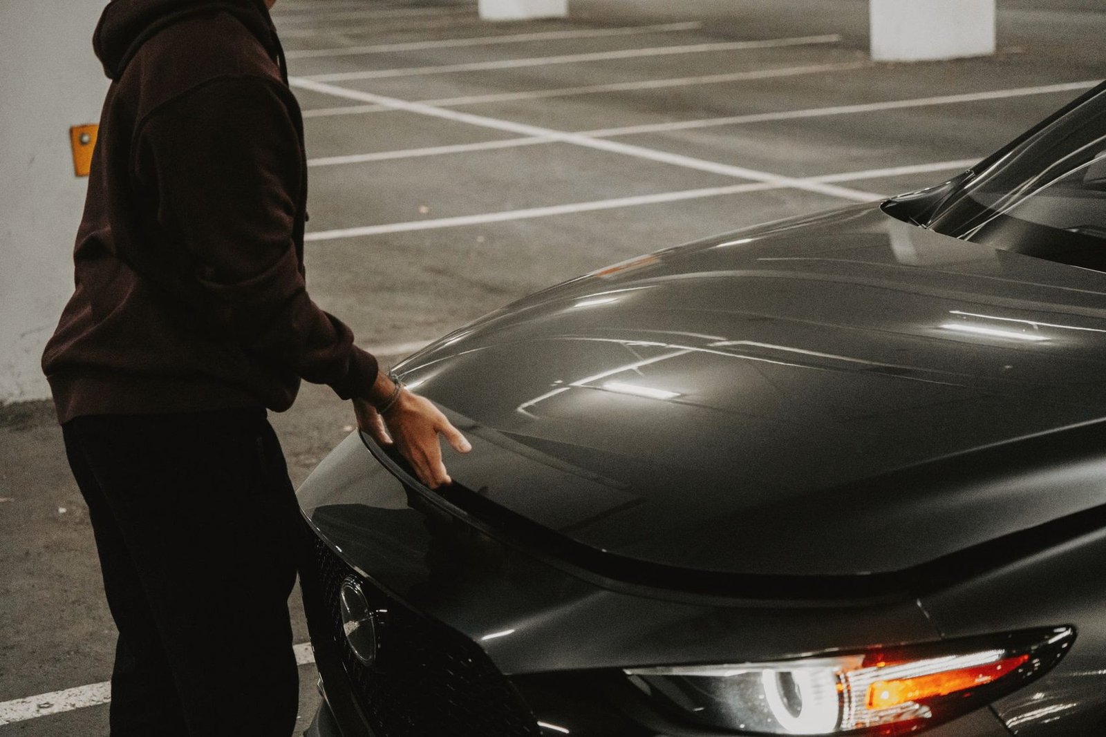 A person in a dark hoodie resting a hand on the hood of a black car in a parking lot