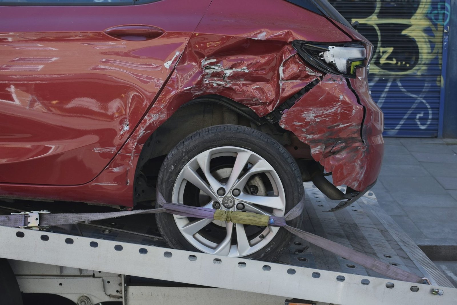 Rear quarter of a red hatchback with crushed bodywork, strapped down by its wheel to a flatbed