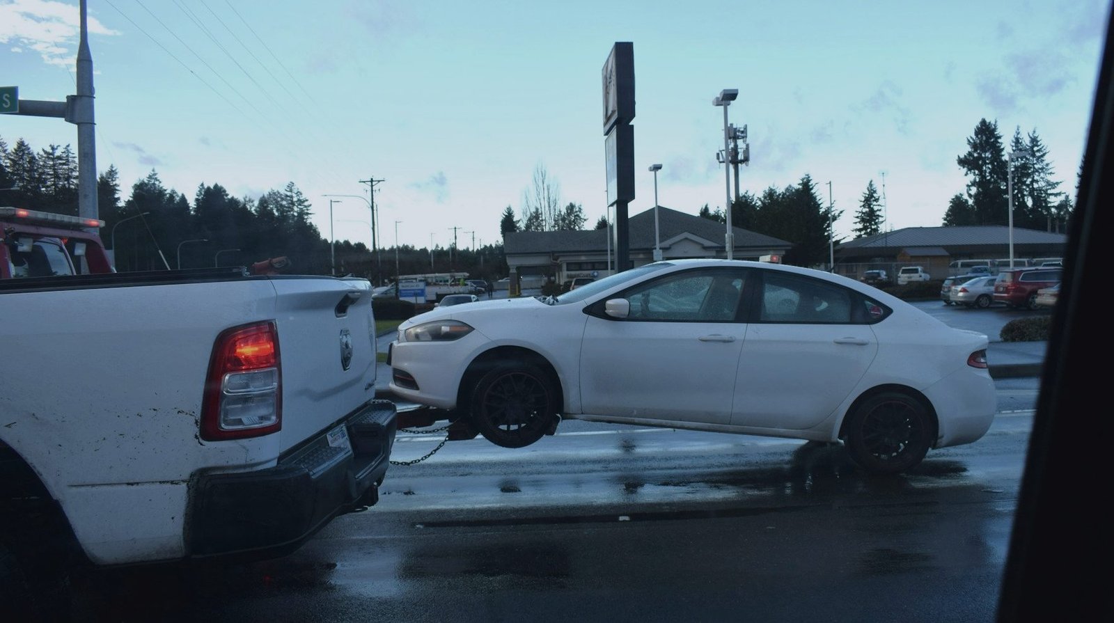 A white sedan with its front wheels lifted, being towed across a wet intersection